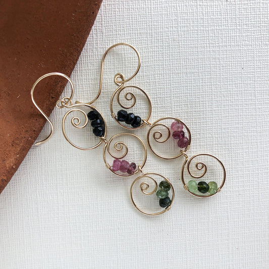 Close-up of multicolor tourmaline dangle earrings – Handmade earrings featuring six gold-filled spiral hoops with wire-wrapped black, green, and pink tourmaline gemstones.