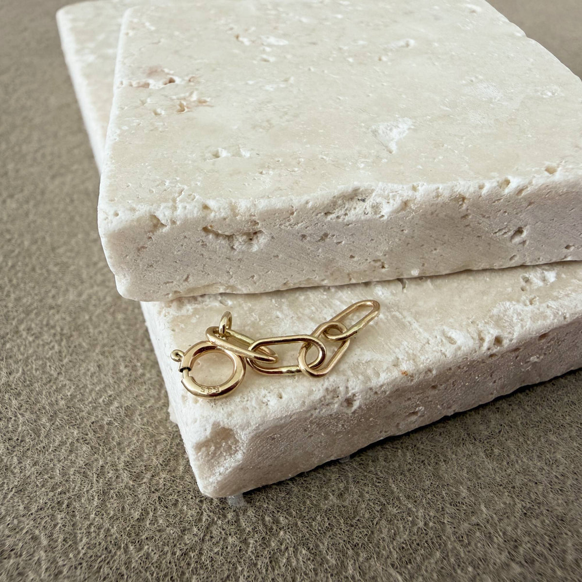 A small 14k handmade solid gold chain extender, placed on top of stacked rectangular cream stone blocks against a textured taupe background.
