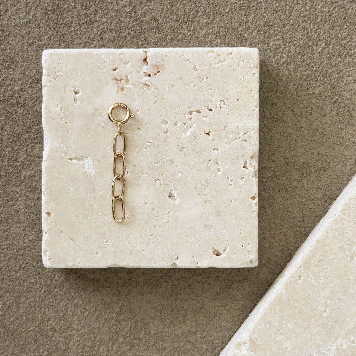 A small 14k gold chain extender with a ring at the top, displayed on a square beige stone tile against a textured taupe background.