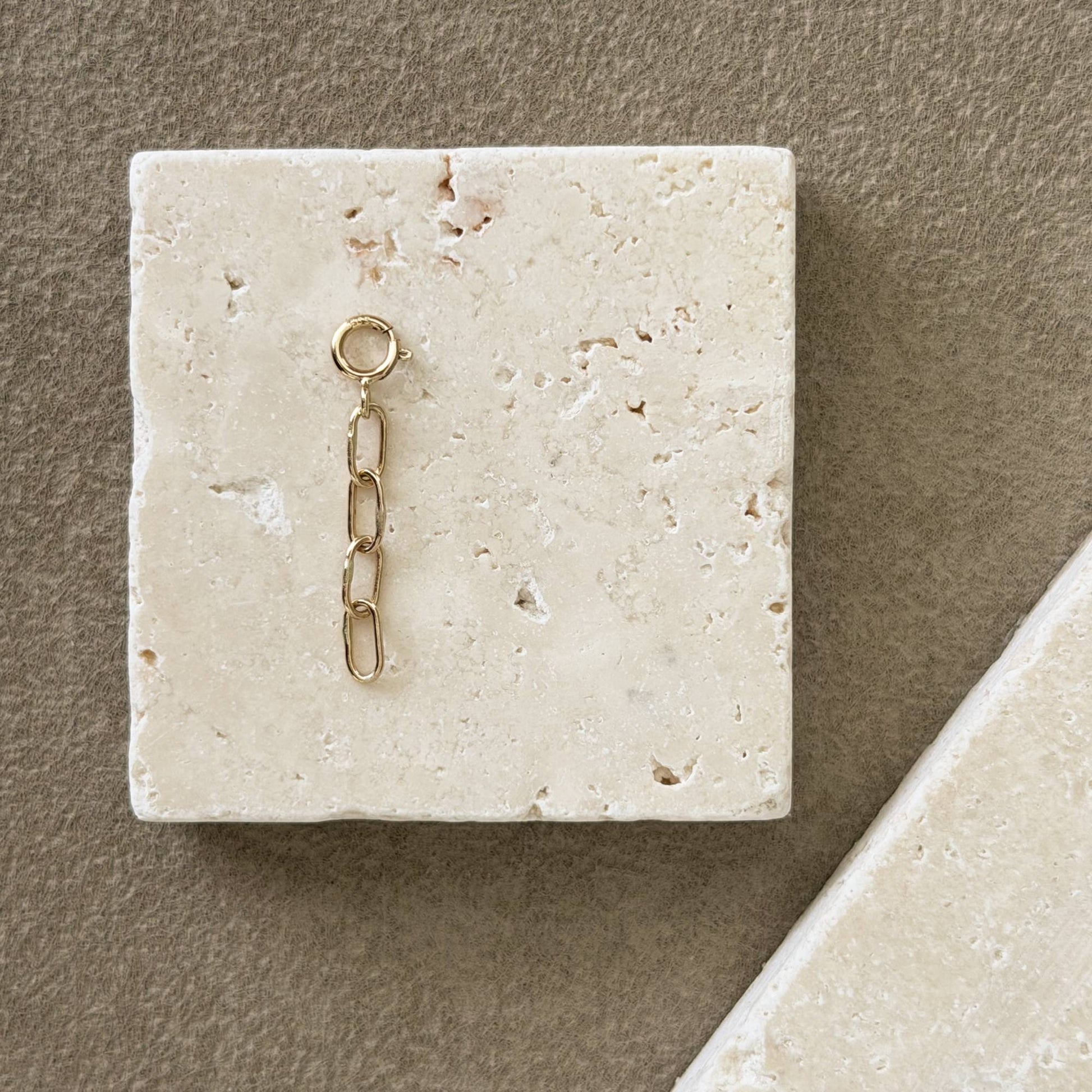 A small 14k gold chain extender with a ring at the top, displayed on a square beige stone tile against a textured taupe background.