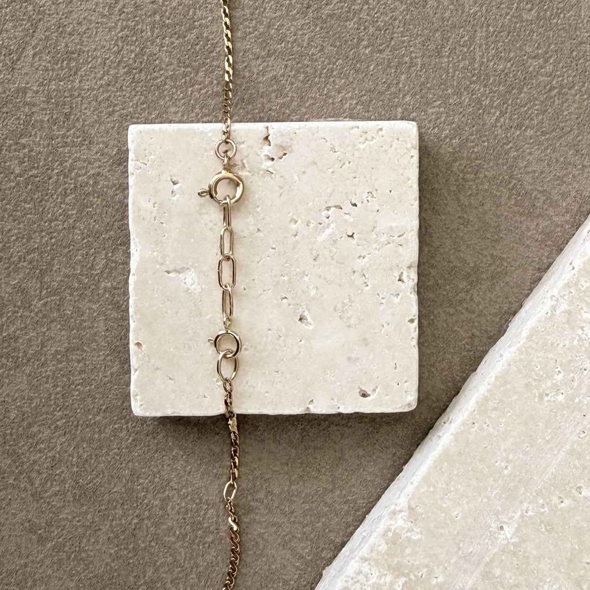 A close-up of a 14k chain necklace with a spring ring clasp and handmade 14k extension chain, draped over a square off-white stone slab against a taupe background.