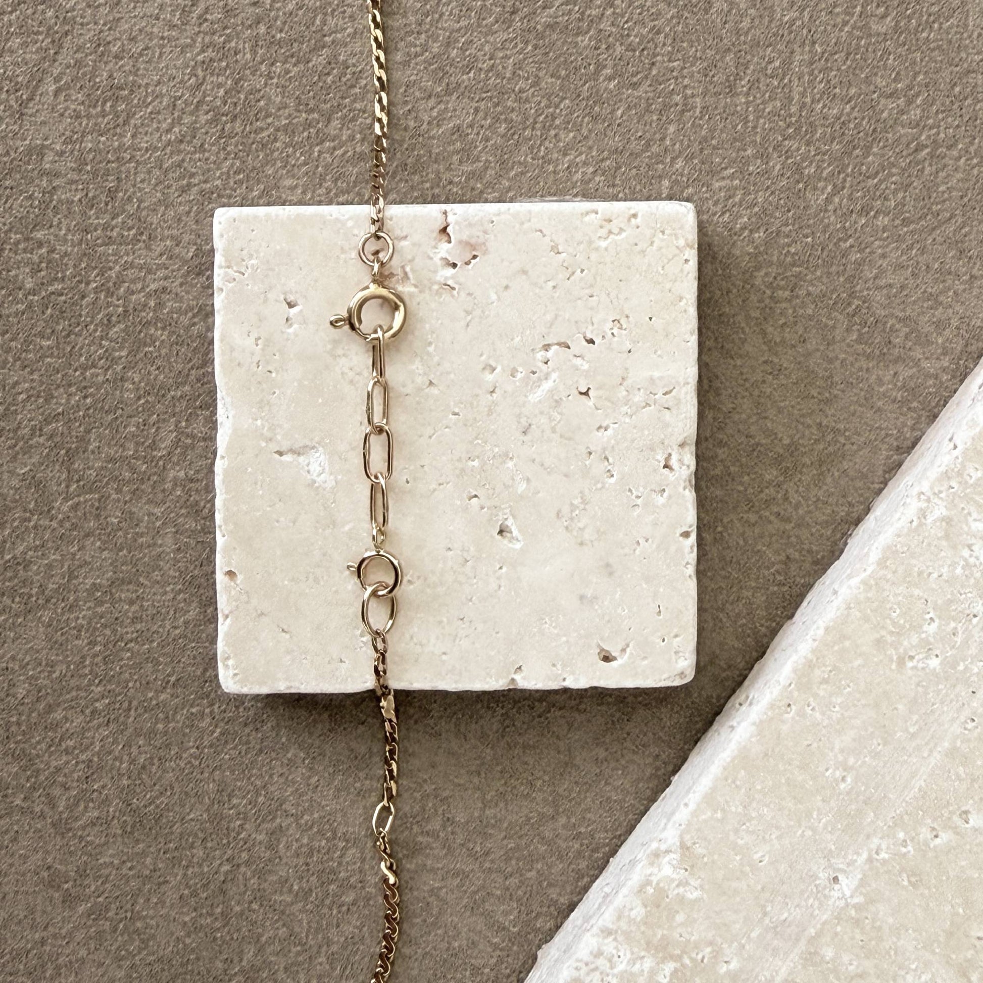 A close-up of a 14k chain necklace with a spring ring clasp and handmade 14k extension chain, draped over a square off-white stone slab against a taupe background.