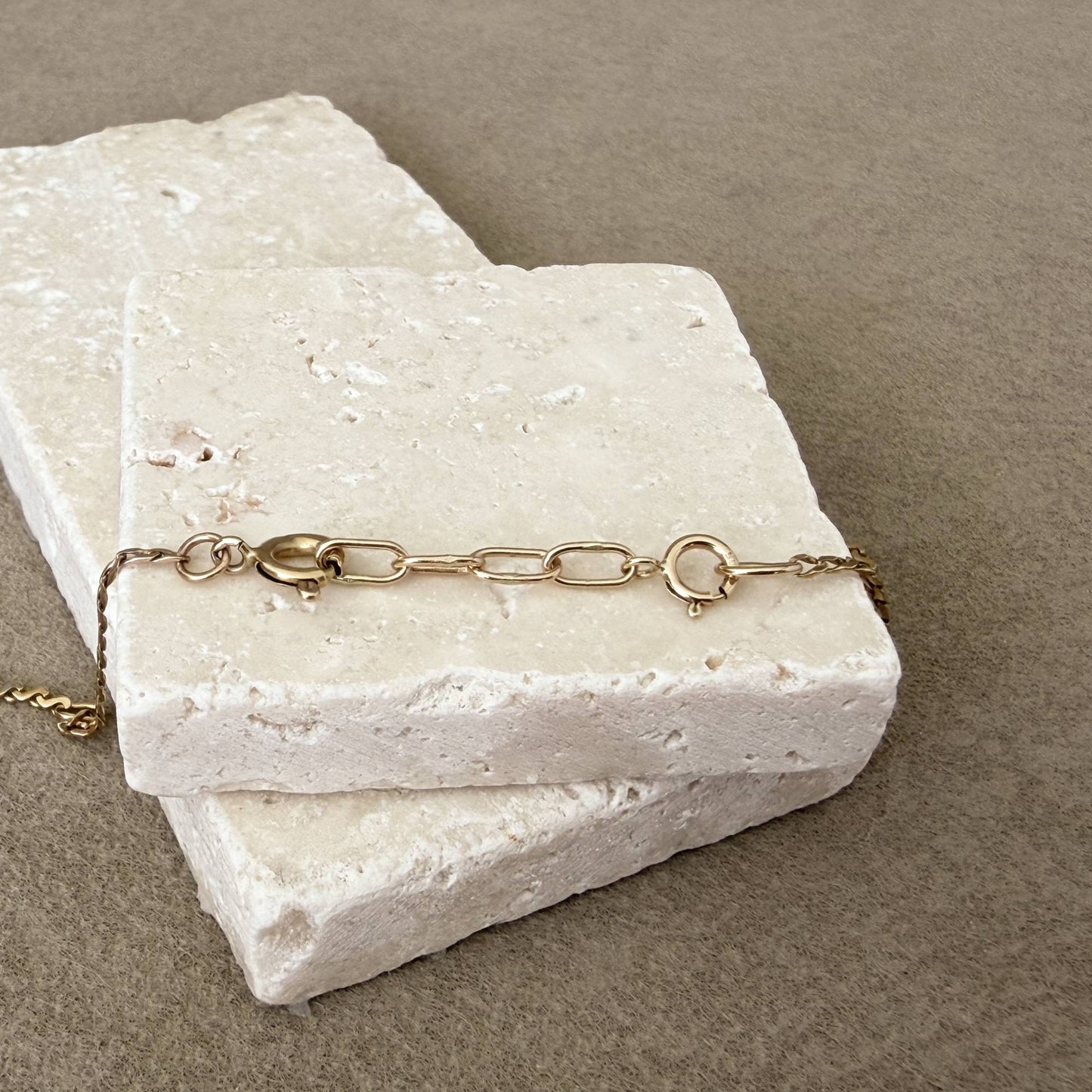 A 14k gold chain with a spring ring clasp and handmade 14k oval links, draped across two stacked rectangular travertine stone blocks on a textured taupe surface.