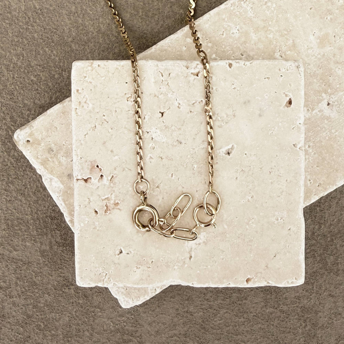 A 14k gold chain necklace with a safety-pin style extender handmade with 14k gold, draped over two stacked beige stone tiles on a textured brown surface.