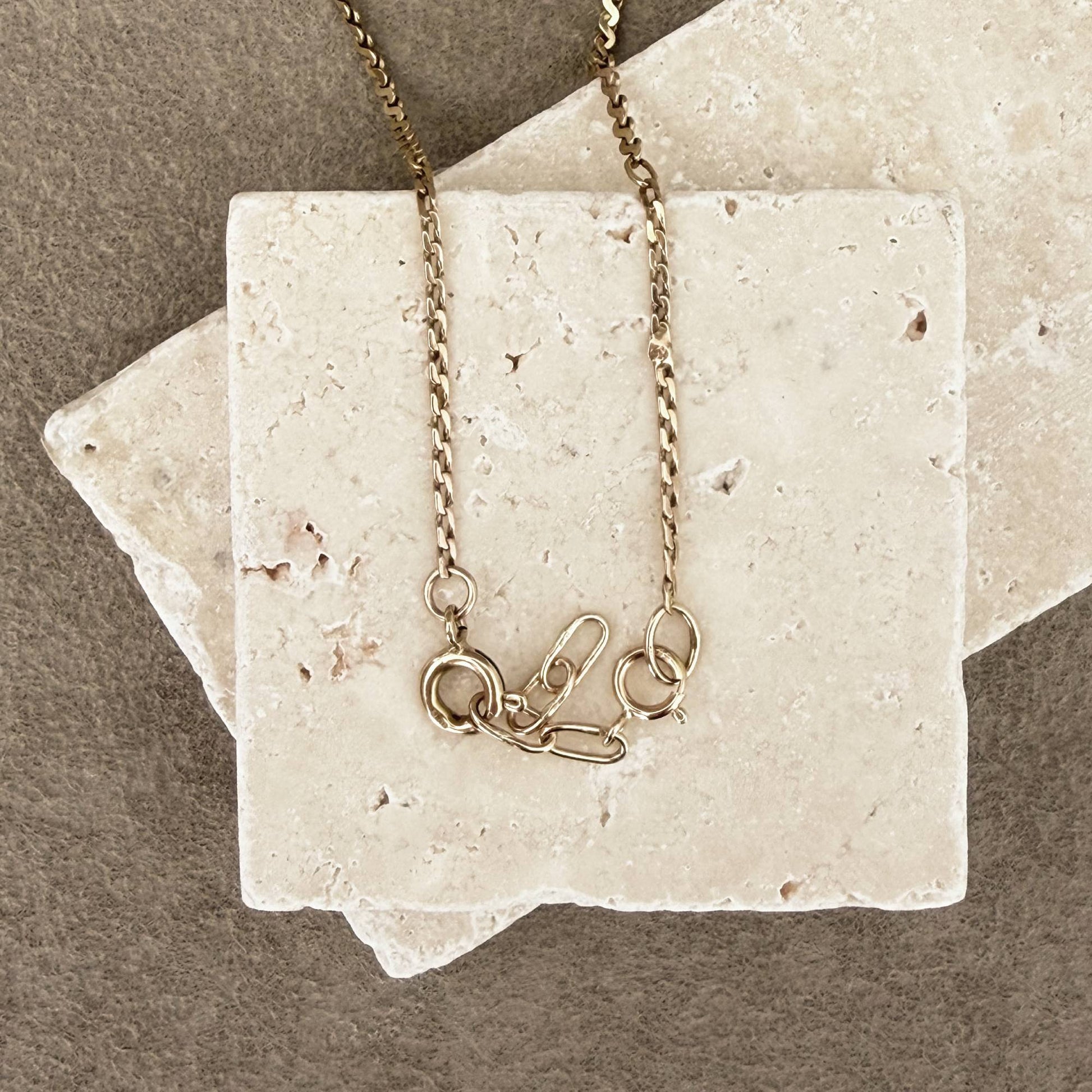 A 14k gold chain necklace with a safety-pin style extender handmade with 14k gold, draped over two stacked beige stone tiles on a textured brown surface.