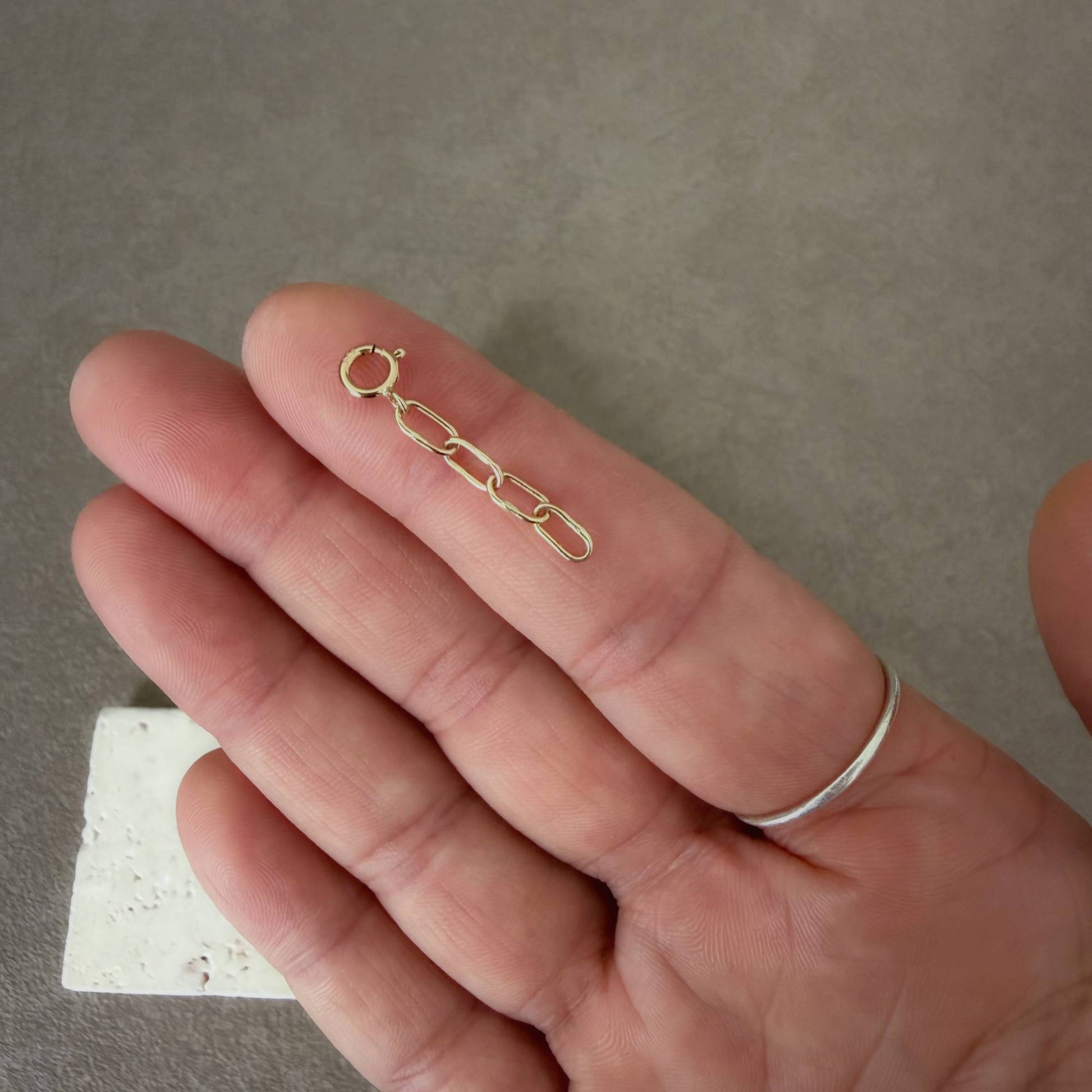 A close-up of a hand holding a small handmade 14k gold chain link extender, with a silver ring visible on the index finger, against a textured gray background.