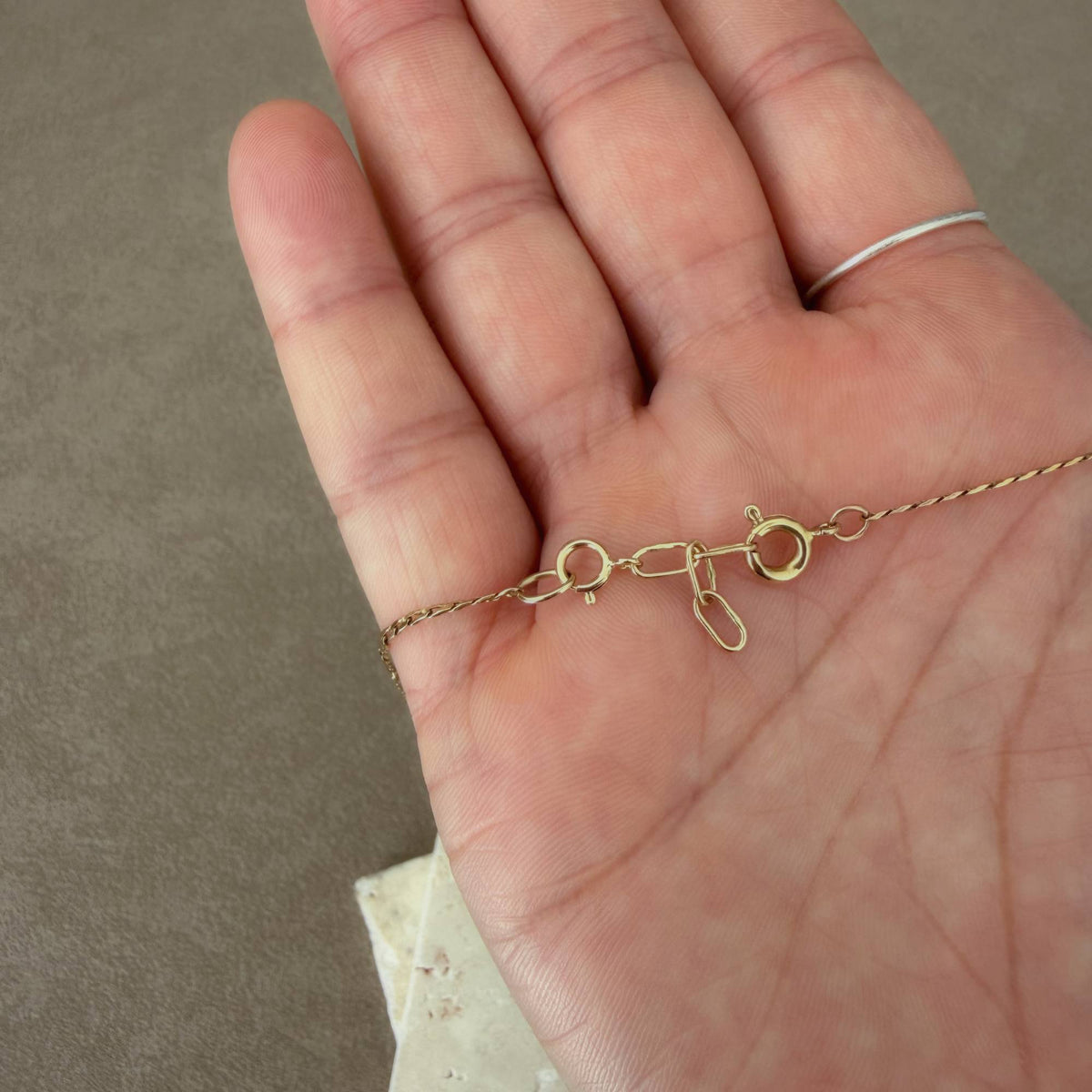 A close-up of a person's hand holding a delicate gold chain with a spring rings clasp and 14k handmade extender, showing the metal details against a neutral gray background.