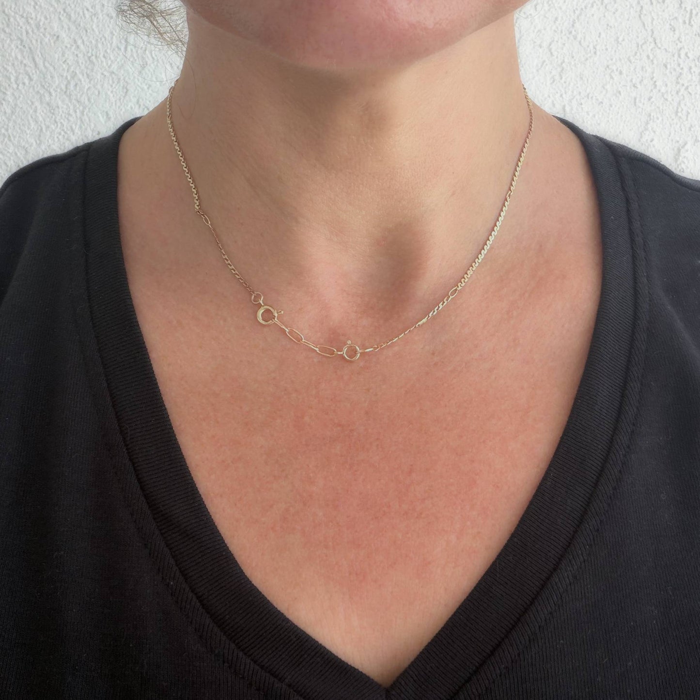 A close-up of a person's neck and chest wearing a delicate gold chain necklace with two small interlocking circular links at the center, styled with a black V-neck top against a light textured wall.