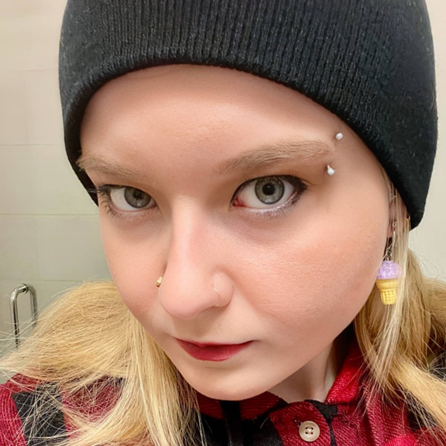 Person wearing a black beanie and red plaid shirt with visible earrings and a gold nose ring
